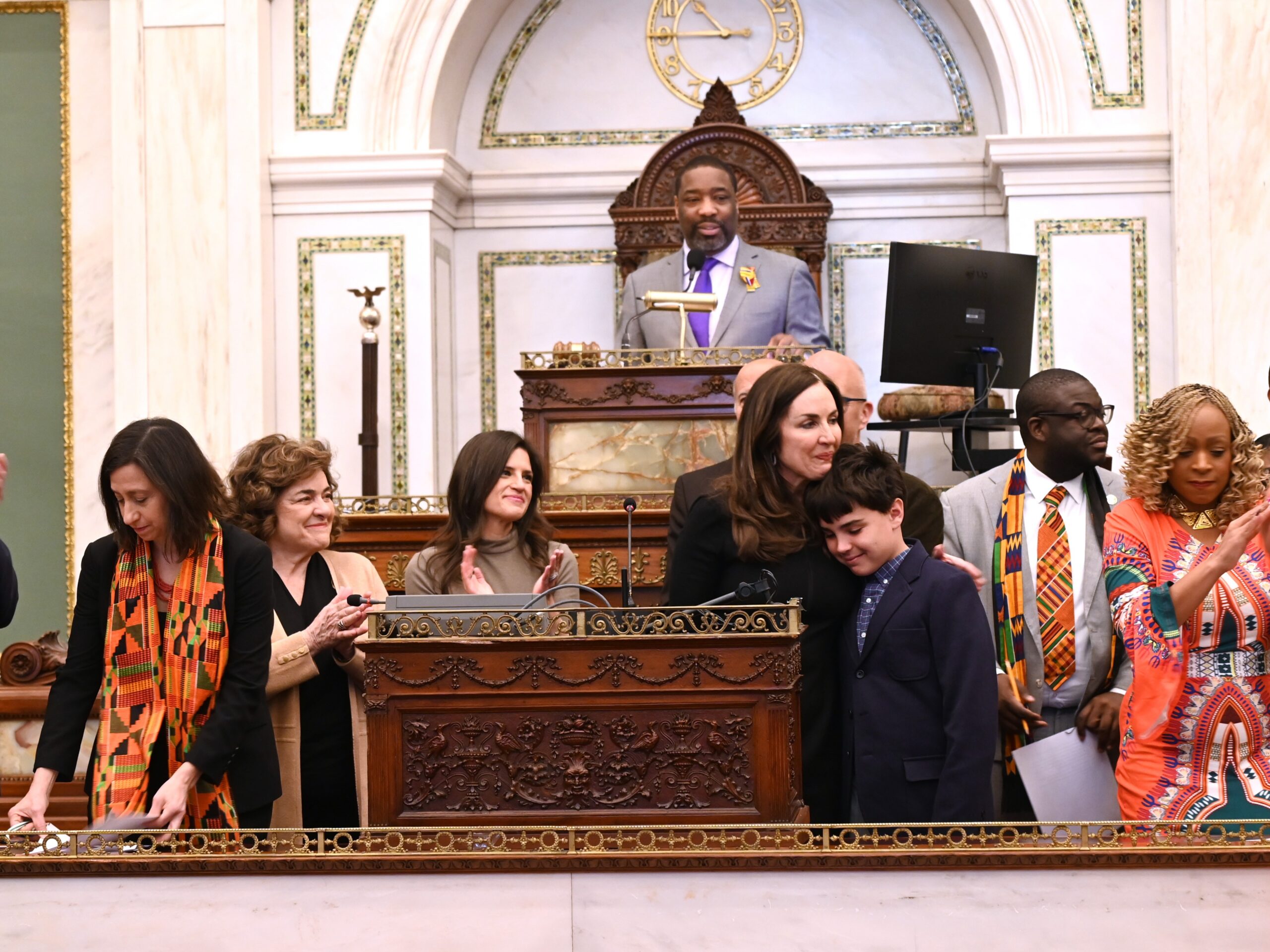 Group gathered at a podium inside Philadelphia City Hall during a ceremony, with several people applauding and a young person standing beside the speaker.