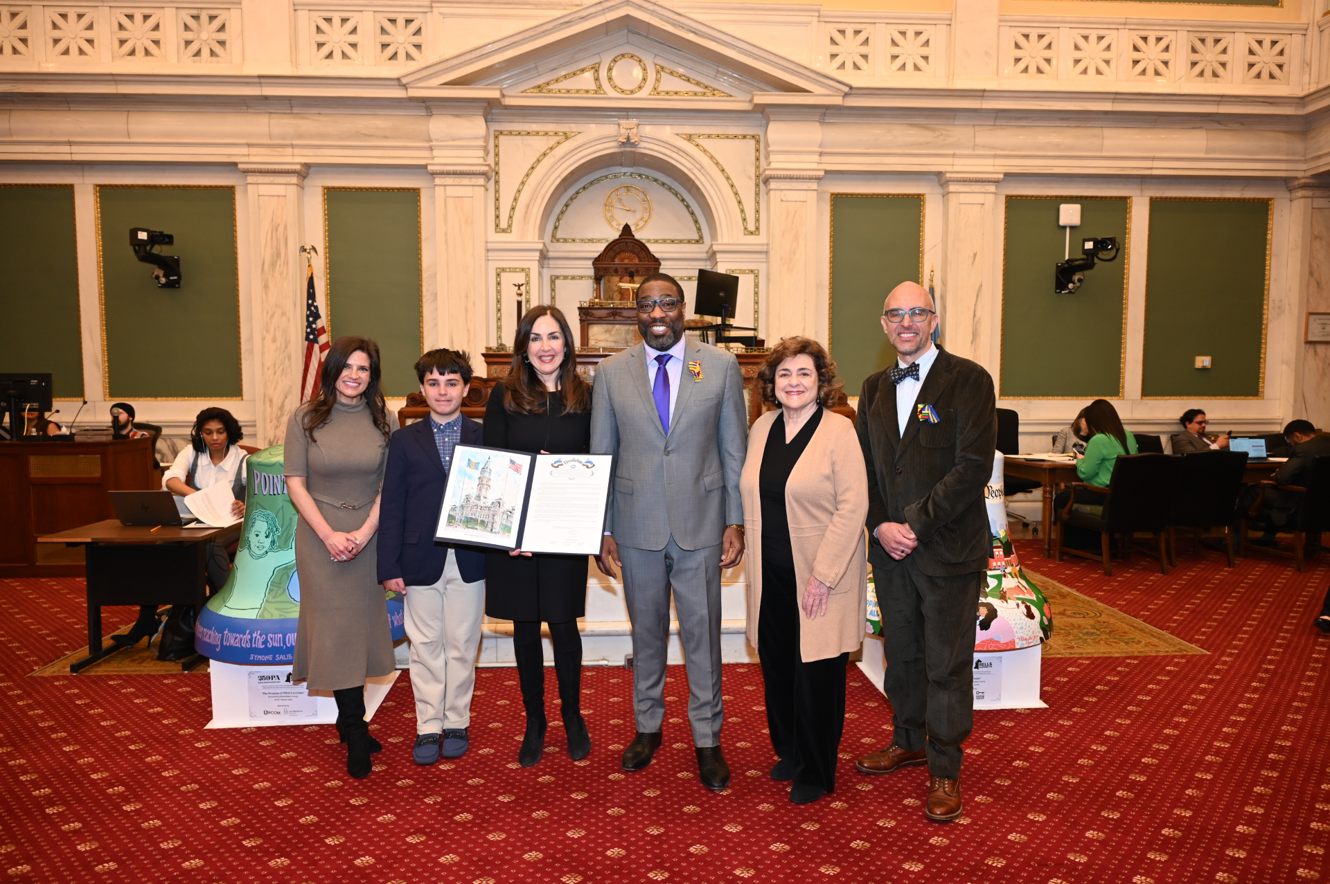 Group of community leaders posing inside Philadelphia City Hall, with a young person and several adults holding a framed proclamation.