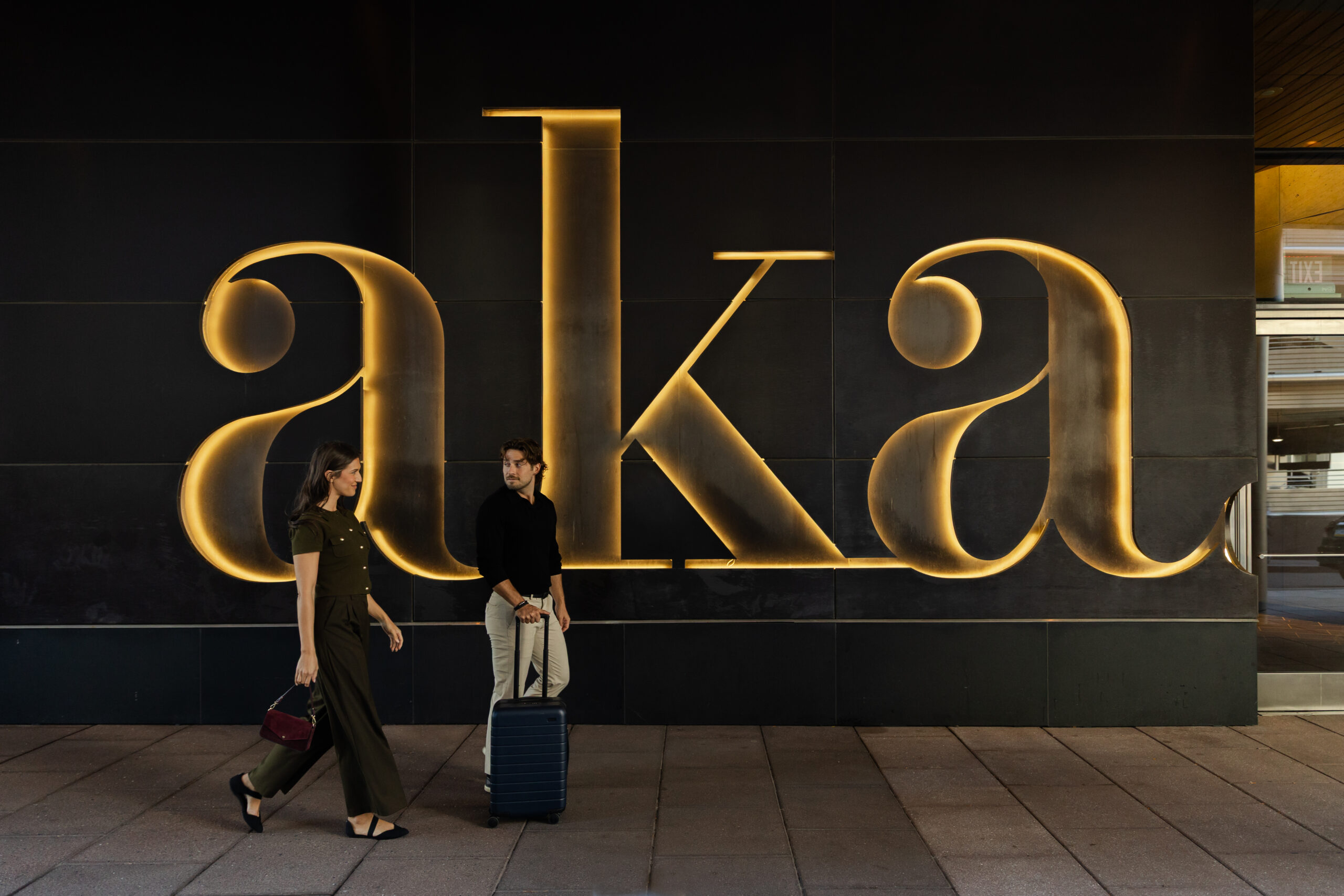 two people walking past a lit up sign for "AKA"